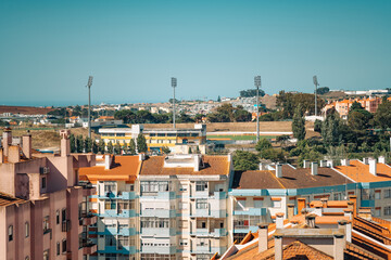 A panoramic view of a residential area with colorful apartment buildings in the foreground and a sports field with floodlights in the background. The image captures a clear, sunny day with blue skies