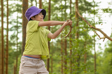 A young boy wearing a green shirt and a purple hat smiles while holding a large stick in a forest, enjoying the natural surroundings and playful activity.