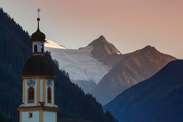The church tower of Neustift im Stubaital at dusk, with the sun setting behind the mountains, a...