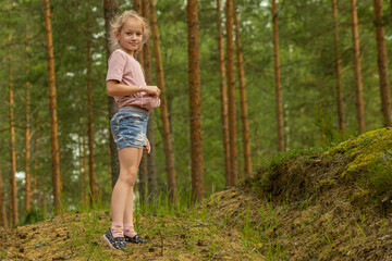 A young girl with blonde hair and wearing a pink shirt and blue floral shorts smiles slightly while standing in a forest.