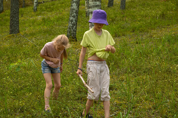 Two children, one in a pink shirt and denim shorts and the other in a green shirt and purple hat, kneel on the forest floor, exploring the natural surroundings.
