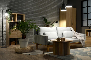 Stylish sofa and wooden table near grey brick wall in interior of living room at night