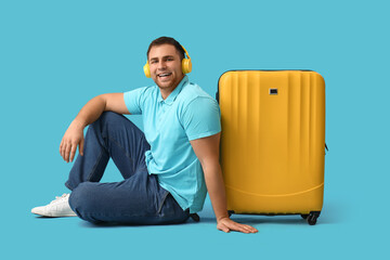 Happy young man with headphones and yellow suitcase on blue background. Travel concept.
