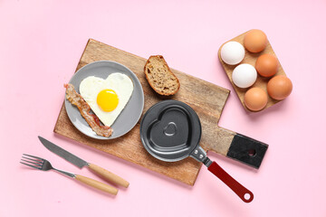 Composition with frying pan, plate with bacon and heart-shaped fried egg on pink background
