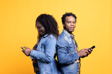 African american spouses posing back to back and browsing social media on their smartphones, looking at videos on online pages. Positive happy couple texting messages in studio.