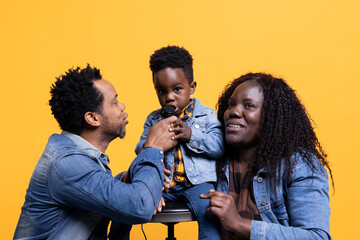 Cheerful adorable small child singing a song on microphone in studio, parents smiling and dancing on his music. Proud mother and father having fun with their happy son on camera.