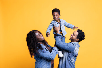 Obraz premium Loving african american parents lifting their cute small son in studio, posing in front of the camera. Proud happy couple bonding with their adorable toddler against yellow background.