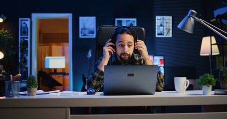 Freelancer doing computer tasks for team project in personal home office while listening music. Remote employee working late at night, typing on laptop keyboard, putting headphones on, camera B