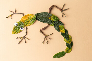 A creative representation of a lizard made using natural materials on a beige paper background. Body: green leaves. Limbs and eyes: smaller leaves and sticks.