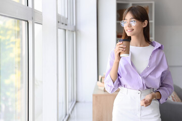 Young woman in eyeglasses with cup of coffee near window at home