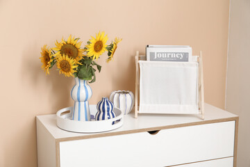 Magazines in holder, tray and vase with beautiful sunflowers on chest of drawers in living room