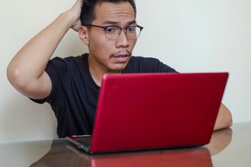 Photo of troubled A Southeast Asian man sit table pc try solve dilemma, wear black t-shirt isolated cream color background,  confused man gesturing with right hand and discussing something.