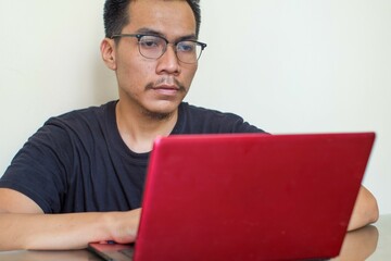 A Southeast Asian Serious man wearing glasses working on laptop online, sitting at table, looking at computer screen, focused male using internet banking service, searching information