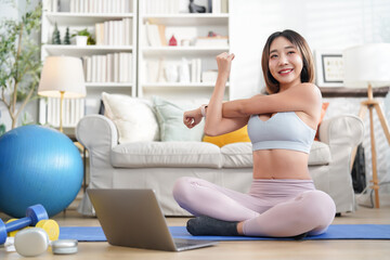 young attractive asian girl in sportswear sitting on yoga mat,warming up exercise,stretching arm with laptop online trainer in the living room