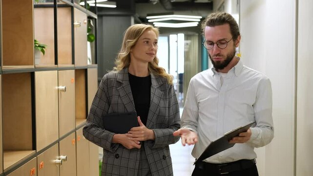 Man walking in a hallway while holding a clipboard when an officemate greet him. Two office workers walk together in a corridor, woman with iPad sharing ideas about his file in clipboard.