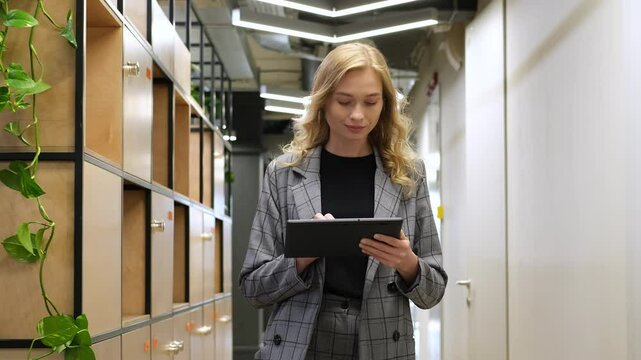 Businesswoman walking in corridor using an iPad. Woman with a smile walks behind the lockers in a modern office.