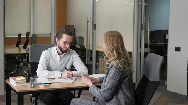 Two professionals engaged in a one-on-one meeting in an office setting. A man with glasses and beard taking notes while a woman gestures as she speaks.