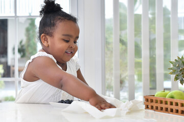Happy little African girl with milk stains on her lips, uses paper to clean up spilled milk on the...