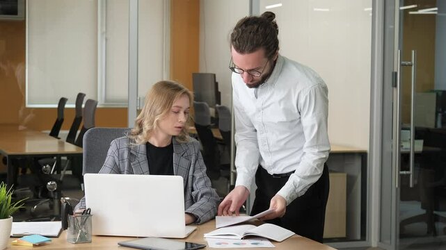 A concentrated businesswoman sitting and working on her laptop. Officemate asking for a signature of his boss on some documents.