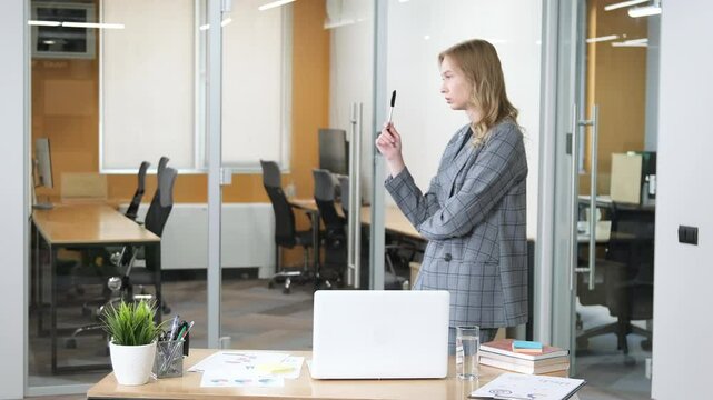 A professional woman in a gray plaid blazer stands in an office, holding a pen to her chin in thought. Female executive planning in office, desk with financial reports and charts.