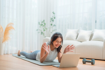Young Asian woman lying on the floor on yoga mat in front of laptop and training at home. Beautiful smiling woman doing stretching exercise while communicates with a trainer online before workout