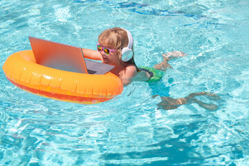 Child sitting in swimming ring in pool and using laptop. Shopping online, freelance concept, summer travel.