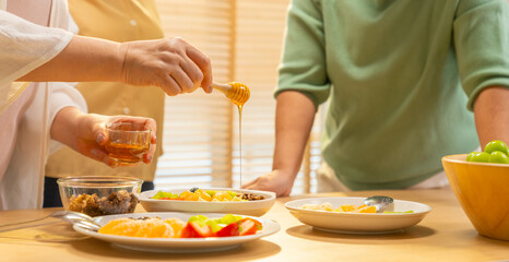 Group of Happy Asian senior women having breakfast together at home. Elderly retired woman friends enjoy healthy lifestyle making homemade greek yogurt with mixed fruit, nuts and honey in the kitchen.