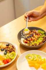 Asian senior woman hand scooping homemade greek yogurt with mixed fruit, nuts and honey on the table. Elderly retired woman enjoy healthy lifestyle cooking and having breakfast in the morning at home.