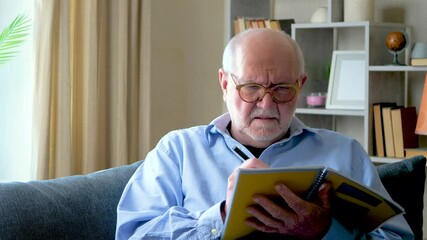 Elderly businessman man in blue shirt sitting on sofa at home doing work remotely checking financial report data. Slow motion. - Powered by Adobe