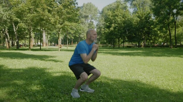 Determined active fitness mature man in sportswear practicing squats exercise, improving core strength and mobility, building stringer leg muscles while working out in summer park.