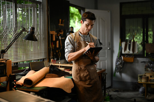 Handsome male artisan wearing an apron using a digital tablet in his workshop. Small business and technology concept
