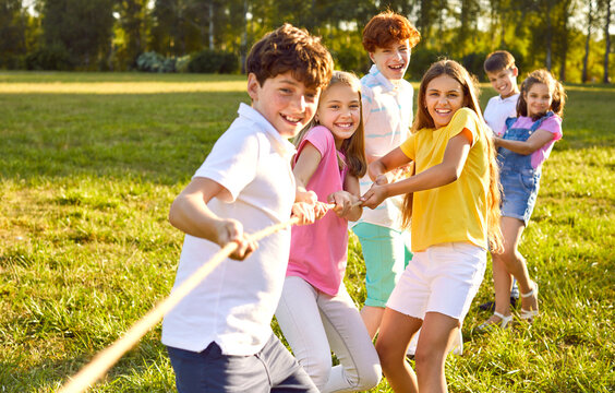Cheerful kids playing fun games on summer day. Group of happy children playing tug of war, standing on green lawn, holding and pulling rope, trying to win victory for their team. Fun, teamwork concept - Powered by Adobe