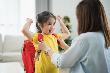A young girl is getting her backpack on with her mother's help. The girl is smiling and seems happy. Back to school concept.