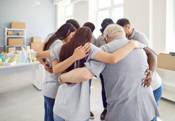 Group of young and mature volunteers standing in a circle hugging and smiling working in community charity center. People volunteering in food bank. Humanitarian aid and social help for poor concept.