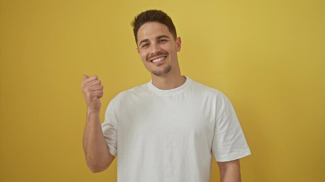 Cheerful young hispanic guy in white t shirt, joyfully pointing and looking to the side against yellow isolated background showing thumbs up