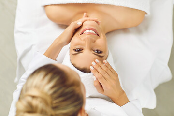 Top view portrait of happy smiling young pretty woman client looking cheerful at camera while lying in spa salon having a face massage and receiving beauty procedure from beautician in beauty clinic.