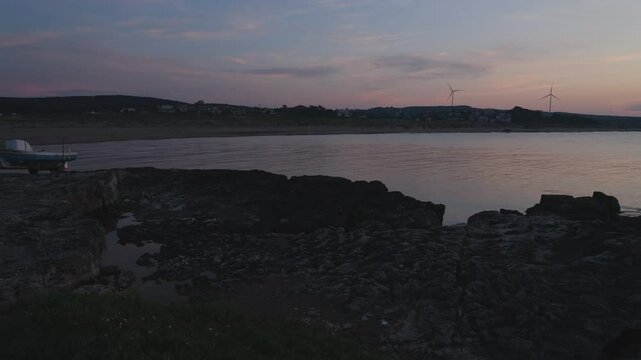 Boat tow tractor on the beach. Evening view and calm sea. Rocky area and wind turbines in the background.