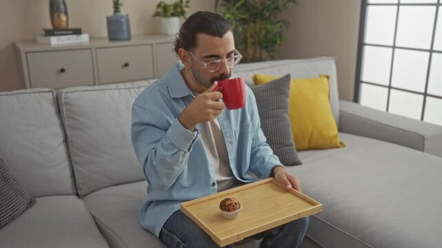 Young man with moustache drinking coffee in cozy home living room while holding a wooden tray with a muffin and sitting on a grey sofa adorned with yellow and grey cushions