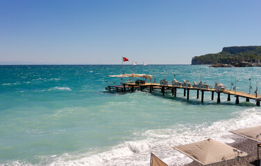 A scenic pier with sun loungers extending into a turquoise sea under a clear blue sky, with a rocky coastline in the background. Concept of relaxation