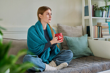 Obraz premium Young woman warming under blanket with mug of hot drink sitting on couch at home