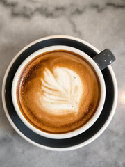 Coffee Latte art with white coffee art flower in brown foam in a black coffee cup on a black saucer on a marble table