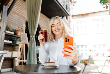 young beautiful blond caucasian woman sitting at table in outdoor terrace of cafe in summer day with chocolate cheesecake and strawberry cocktail, businesswoman looking at smartphone and smiling