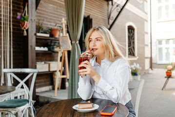 young beautiful blond caucasian woman sitting at table in outdoor terrace of cafe in summer day with chocolate cheesecake, drinking strawberry cocktail