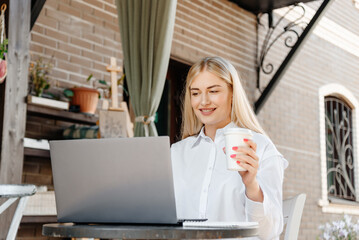 young beautiful blond caucasian woman sitting at table in outdoor terrace of cafe in summer day with notebook, businesswoman working, freelancer holding paper glass of coffee and smiling