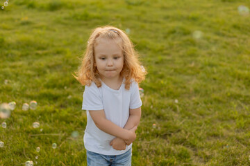 Fototapeta premium Cute blonde kid with curly hair walks on the lawn with soap bubbles in summer