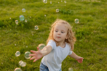 Cute blonde kid with curly hair walks on the lawn with soap bubbles in summer