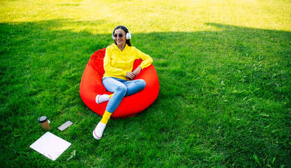 Fototapeta premium Cute woman relaxing on bean bag chair sitting and resting, using laptop, happy millennial girl leaning back, enjoying job, feeling pleased and satisfied. Female outdoors on lawn in park or garden