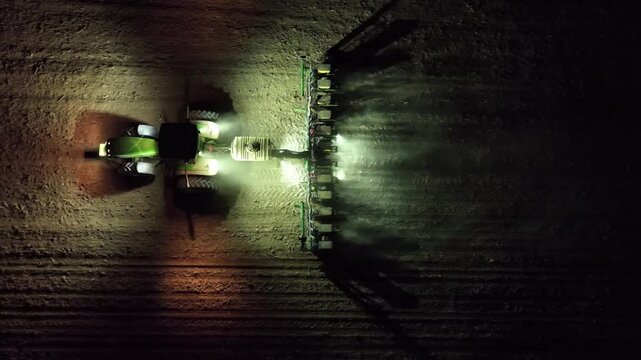 arieal high-angle drone footage of an agricultural tractor planting seeds in a field at night. The tractor's powerful lights illuminate the field, advanced planting techniques and nighttime agricultur