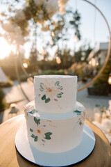 White wedding cake in 2 tiers on a white stand. The cake is decorated with chamomile flowers and green leaves. Cake on the street in the evening at sunset. Glare of the sun. Blurred background