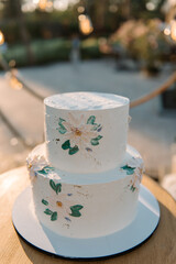 White wedding cake in 2 tiers on a white stand. The cake is decorated with chamomile flowers and green leaves. Cake on the street in the evening at sunset. Glare of the sun. Blurred background